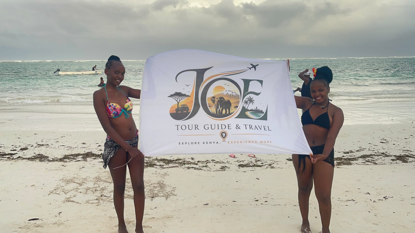 Two guests holding the JOE Tour Guide & Travel flag on a Mombasa beach